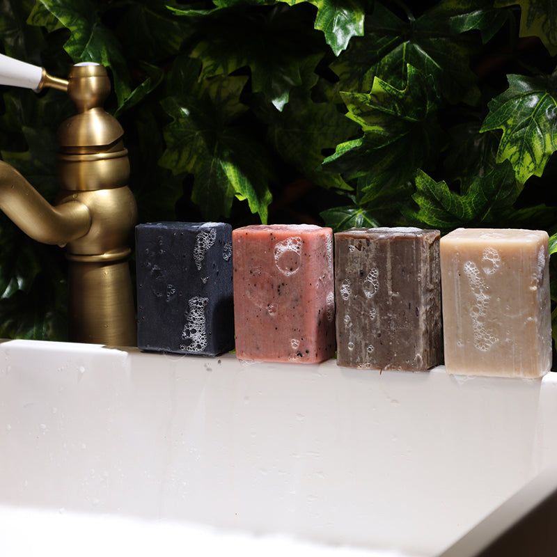 Four soap bars of different colors on a white surface with a green leafy background